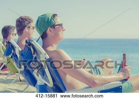 Stock Image - Young men drinking beer in deck chairs on sunny beach. Fotosearch