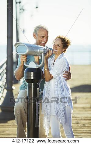 Stock Image - Couple using coin-operated binoculars on pier. Fotosearch