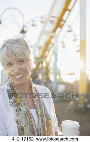 Stock Image - Portrait of smiling senior woman drinking coffee at amusement park. Fotosearch