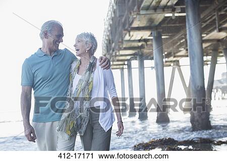 Stock Image - Senior couple laughing near pier at beach. Fotosearch