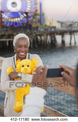 Stock Image - Senior woman with teddy bear posing for photograph at amusement park. Fotosearch