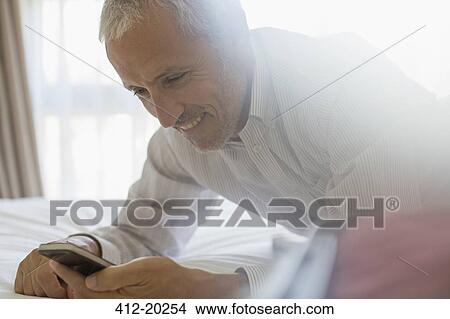 Picture - Businessman using cell phone on bed in hotel room. Fotosearch