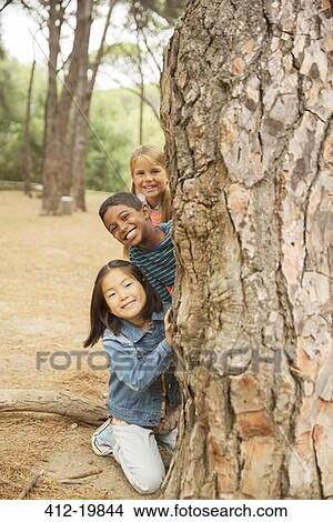 Picture - Children peeking out from behind tree in forest. Fotosearch