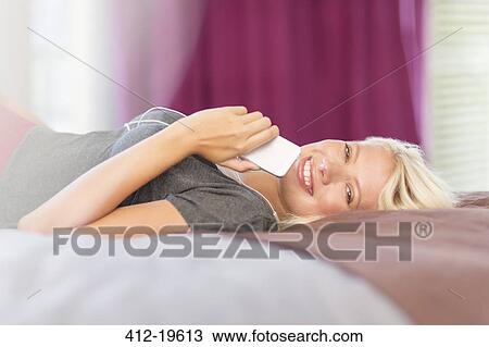 Stock Image - Woman holding cell phone on bed. Fotosearch