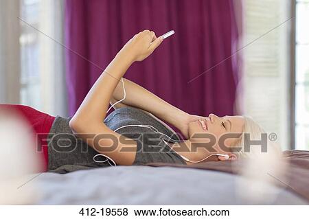 Woman listening to earbuds on bed View Large Photo Image Stock Photo - Woman listening to earbuds on bed. Fotosearch