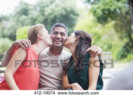 Picture - Women kissing man's cheeks outdoors. Fotosearch