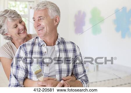 Stock Image - Older couple hugging with paintbrush in living space. Fotosearch