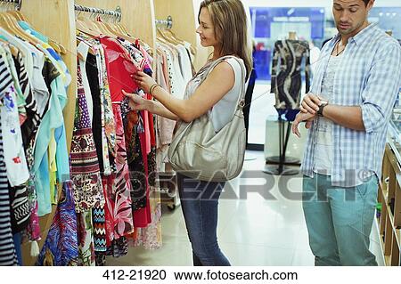 Stock Image - Bored man shopping with girlfriend in clothing store. Fotosearch