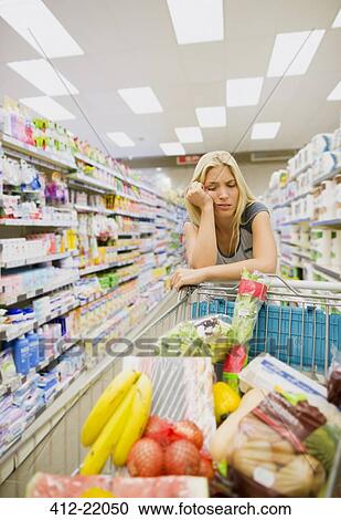Stock Image - Bored woman pushing full shopping cart in grocery store. Fotosearch