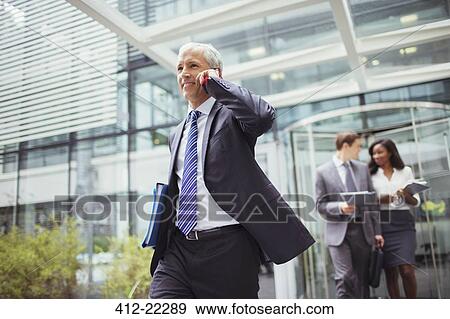 Businessman talking on phone while walking out of office building View Large Photo Image Stock Photo - Businessman talking on phone while walking out of office building. Fotosearch