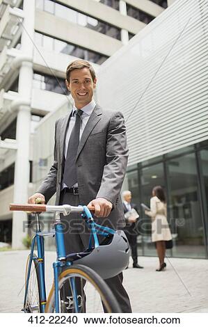Stock Image - Businessman walking bicycle outside of office building. Fotosearch