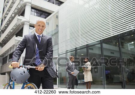 Stock Image - Businessman walking bicycle outside of office building. Fotosearch