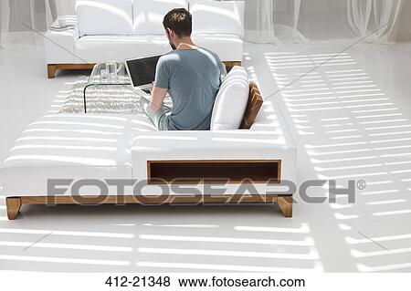 Stock Photo - Man using laptop on sofa in modern living room. Fotosearch