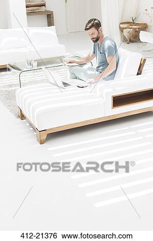 Man using laptop on sofa in modern living room View Large Photo Image Stock Photograph - Man using laptop on sofa in modern living room. Fotosearch