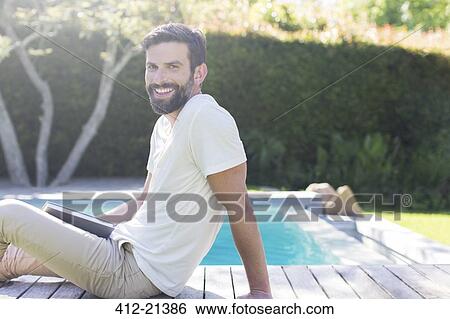 Stock Photograph - Smiling man relaxing on wooden deck by swimming pool. Fotosearch