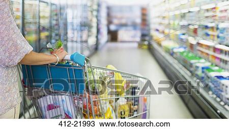 Stock Image - Woman pushing full shopping cart in grocery store. Fotosearch