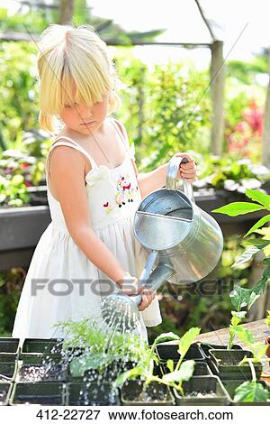 Stock Photo - Blond girl wearing sundress watering potted seedlings in greenhouse. Fotosearch