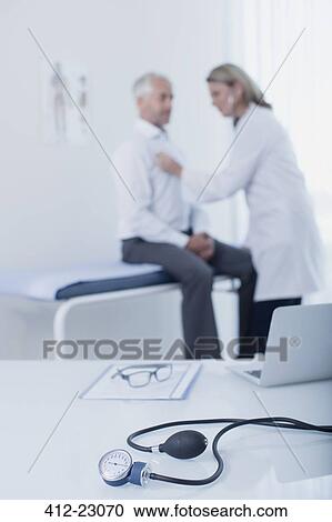 Blood pressure gauge, laptop, file and glasses on desk in doctor's office, female doctor examining patient in background View Large Photo Image Stock Image - Blood pressure gauge, laptop, file and glasses on desk in doctor's office, female doctor examining patient in background. Fotosearch