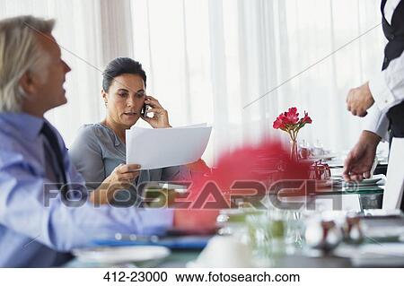 Stock Image - Businesswoman looking at chart and talking on mobile phone at restaurant table. Fotosearch