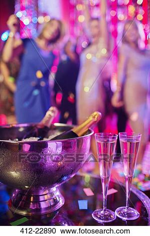 Stock Image - Champagne bottles in ice bucket and champagne flutes in nightclub, people dancing in background. Fotosearch