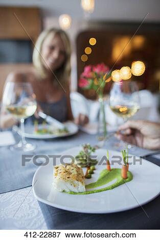 Stock Photo - Close up of fancy plate with fish and carrots on restaurant table, woman in background. Fotosearch