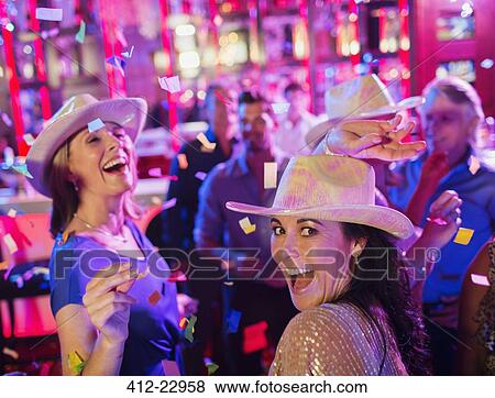 Stock Photo - Confetti falling on women wearing cowboy hats laughing and dancing in nightclub. Fotosearch