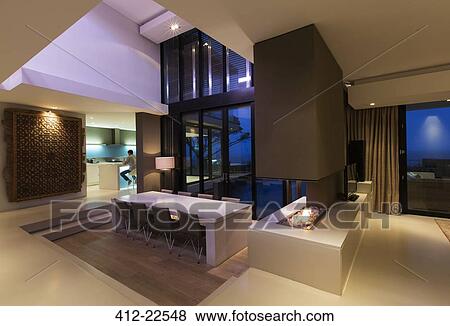 Stock Photo - Dining area in modern house at night, man in kitchen in background. Fotosearch
