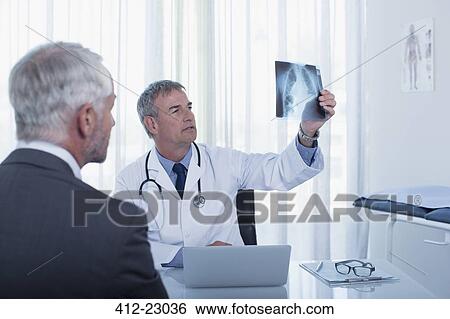 Stock Photograph - Doctor sitting at desk in office examining patient's x-ray. Fotosearch