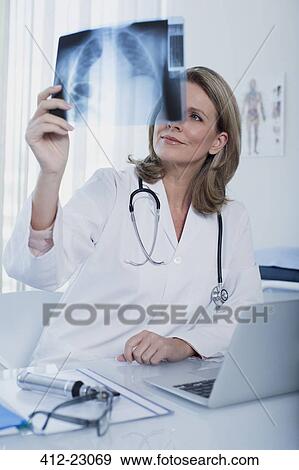 Stock Photo - Female doctor looking at x-ray at desk with laptop in office. Fotosearch