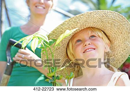 Girl wearing straw hat holding plant, woman with shovel in background View Large Photo Image Stock Image - Girl wearing straw hat holding plant, woman with shovel in background. Fotosearch