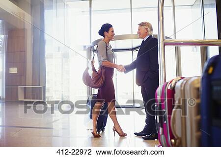 Stock Image - Man and woman shaking hands in hotel lobby, luggage cart in foreground. Fotosearch