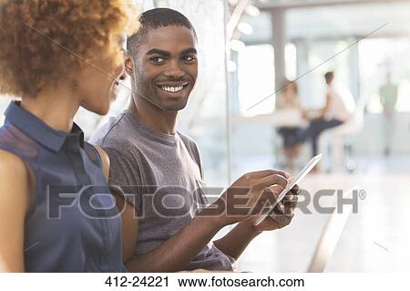 Stock Image - Man and woman using tablet in office, colleagues in background. Fotosearch