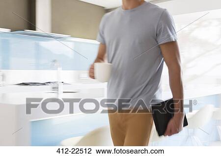 Stock Image - Man carrying coffee cup and digital tablet walking through modern kitchen. Fotosearch