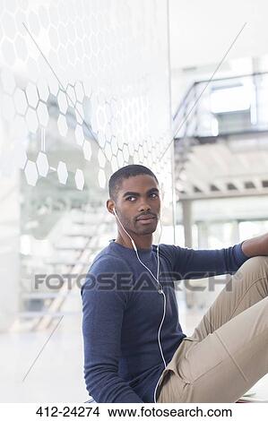 Picture - Man listening to music on earphones, sitting on floor in office corridor. Fotosearch