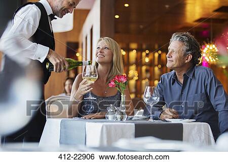 Stock Photograph - Mature couple sitting at restaurant table, waiter pouring white wine into woman's glass. Fotosearch