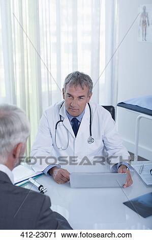 Mature doctor with laptop and man sitting at desk in office View Large Photo Image Stock Image - Mature doctor with laptop and man sitting at desk in office. Fotosearch