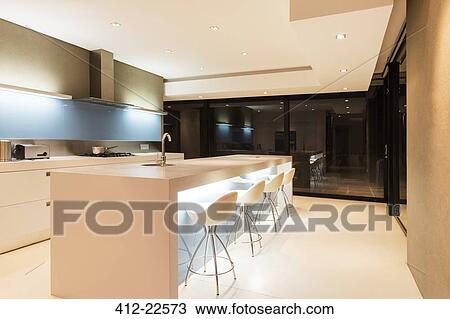 Stock Image - Modern white kitchen with kitchen island and stools illuminated at night. Fotosearch