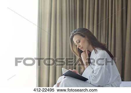Stock Photography - Pensive woman wearing white bathrobe writing in notepad in bedroom. Fotosearch