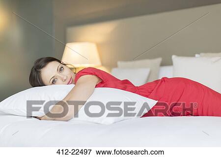 Stock Photo - Portrait of beautiful woman wearing red dress lying on bed and hugging pillow. Fotosearch
