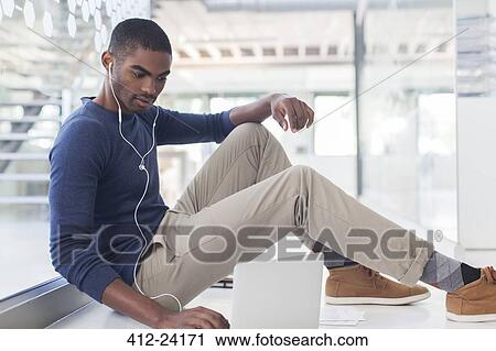 Stock Image - Portrait of businessman sitting on floor in modern office, using laptop and earphones. Fotosearch