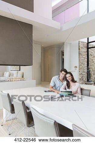 Stock Photograph - Portrait of couple sitting in dining room with books, newspaper and digital tablet on table. Fotosearch