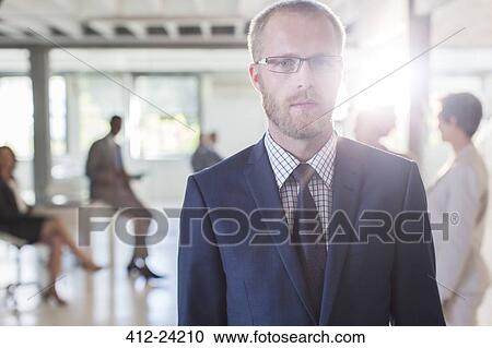 Stock Image - Portrait of serious businessman wearing glasses and suit standing in office, colleagues in background. Fotosearch