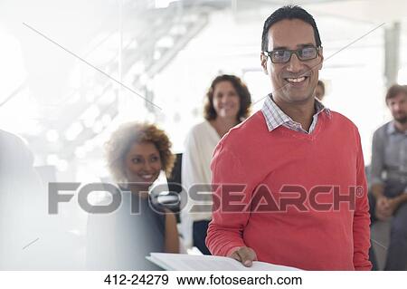 Stock Photo - Portrait of smiling businessman wearing glasses and pink sweatshirt with team in background. Fotosearch