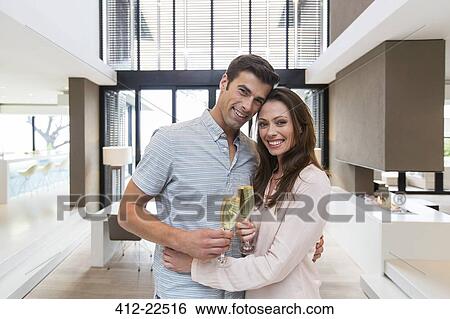 Portrait of smiling couple embracing and holding champagne flutes in modern living room View Large Photo Image Stock Photograph - Portrait of smiling couple embracing and holding champagne flutes in modern living room. Fotosearch