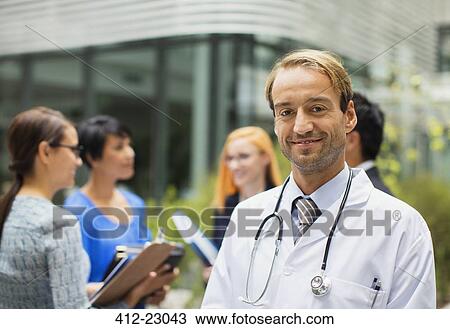 Stock Image - Portrait of smiling doctor wearing lab coat standing in front of hospital, women with clipboards in background. Fotosearch