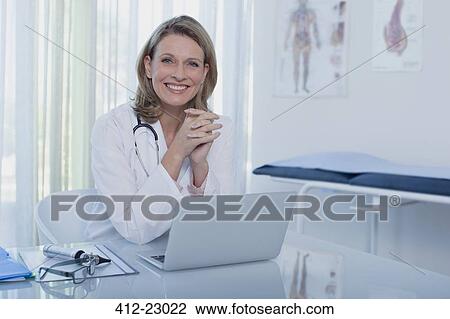 Stock Image - Portrait of smiling female doctor sitting at desk with laptop. Fotosearch