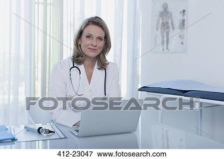 Portrait of smiling female doctor sitting at desk with laptop in office View Large Photo Image Stock Photo - Portrait of smiling female doctor sitting at desk with laptop in office. Fotosearch