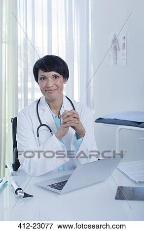 Portrait of smiling female doctor sitting at desk with laptop in office View Large Photo Image Stock Photo - Portrait of smiling female doctor sitting at desk with laptop in office. Fotosearch