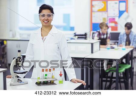 Portrait of smiling female teacher wearing protective eyewear, standing behind desk with microscope and test tubes in rack, students in background View Large Photo Image Stock Image - Portrait of smiling female teacher wearing protective eyewear, standing behind desk with microscope and test tubes in rack, students in background. Fotosearch