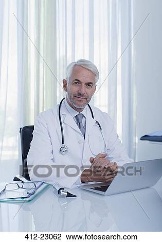 Portrait of smiling mature doctor sitting at desk with laptop in office View Large Photo Image Stock Image - Portrait of smiling mature doctor sitting at desk with laptop in office. Fotosearch